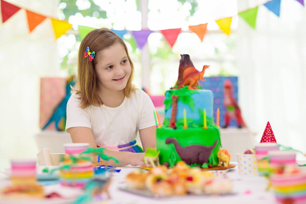 Çocukların doğum günü partisi. Dinozor temalı pasta. Küçük kız mumları üflüyor ve hediyeleri açıyor. Çocuk etkinliği. Dinazorlar için dekorasyon temalı kutlama. - Fotoğraf, Görsel