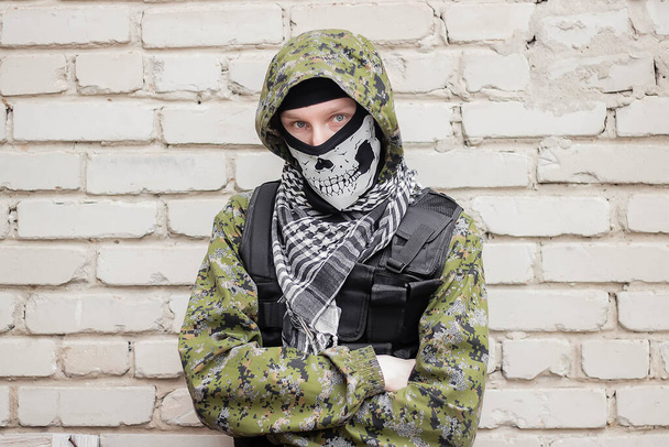 A man in camouflage, a balaclava with a skull and a bulletproof vest. against the background of a brick wall. A criminal, a gangster is preparing an attack from the corner. - Foto, Bild