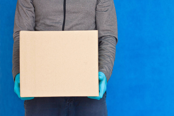 Courier hand holds a brown box on a blue background in rubber gloves on a face mask. Detail of a man carrying a cardboard parcel with copy space - Fotoğraf, Görsel
