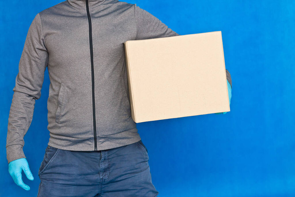 Courier hand holds a brown box on a blue background in rubber gloves on a face mask. Detail of a man carrying a cardboard parcel with copy space - Photo, Image