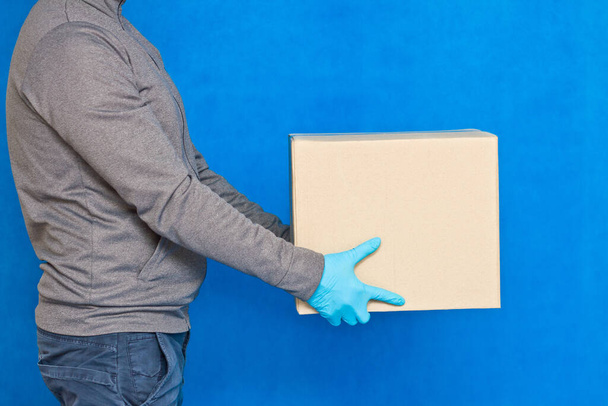 La mano del mensajero sostiene una caja marrón sobre un fondo azul con guantes de goma sobre una mascarilla. Detalle de un hombre que lleva un paquete de cartón con espacio para copias
 - Foto, Imagen