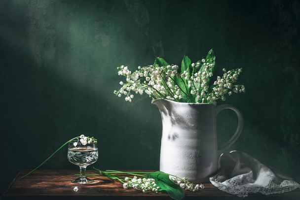 Classic still life with beautiful white Lily of the valley bouquet in white jar and a glass of water in ray of light. Art photography. - Photo, Image