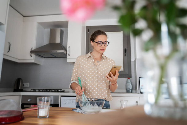 Young happy charming woman girl with a kitchen preparing dessert and watching a recipe in a smartphone on the Internet, cooking at home, hobbies and entertainment. - Foto, Bild