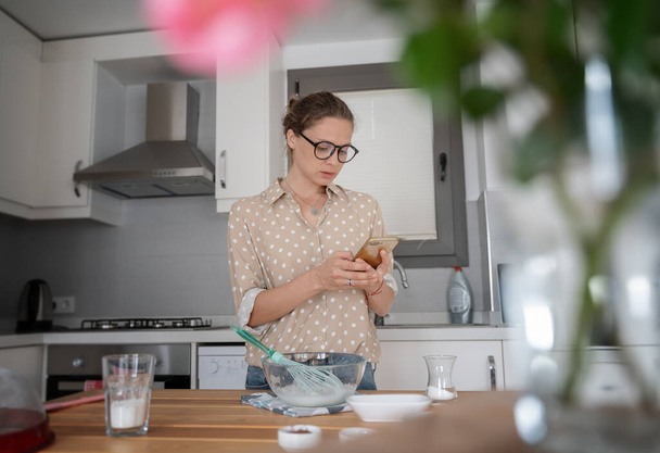 Giovane ragazza donna affascinante felice con una cucina preparare il dessert e guardare una ricetta in uno smartphone su Internet, cucinare a casa, hobby e intrattenimento
. - Foto, immagini