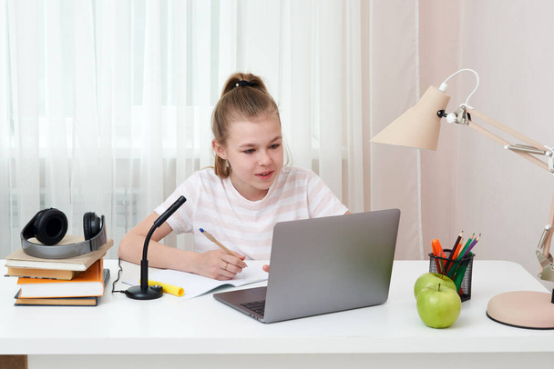 İnternette kulaklık ve dizüstü bilgisayarla öğrenen genç bir kızın portresi. Evdeki masasında oturmuş ödev yaparken not alıyor. - Fotoğraf, Görsel