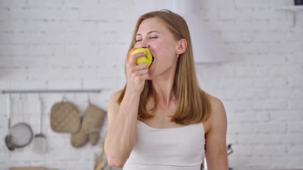 Fit sonriente mujer joven comiendo fruta sana. Una guapa chica disfruta comiendo manzana dulce fresca en el fondo de la cocina moderna. Concepto de pérdida de peso. - Metraje, vídeo