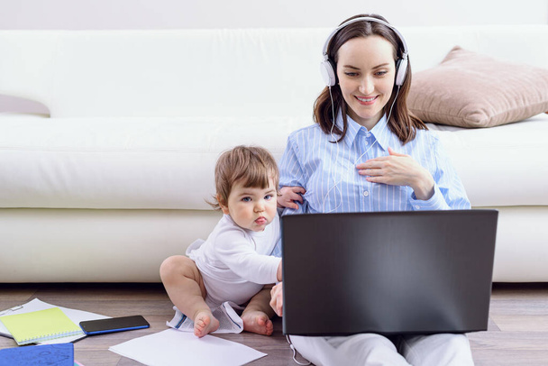 Frau mit Kopfhörer blickt auf Laptop-Bildschirm, Kind sitzt neben ihr, Lernkonzept zu Hause, Fernarbeit zu Hause - Foto, Bild