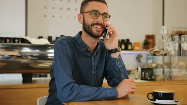 Young happy attractive man in the glasses talking on the smart phone while sitting at the table with cup of coffee in the cafe. Portrait shot. - Imágenes, Vídeo