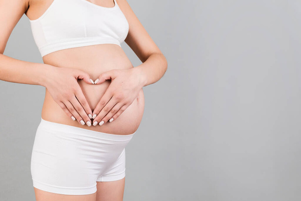 Close up of young pregnant woman making heart shape with her hands on the belly at gray background. Love to the future baby. Copy space. - Photo, Image