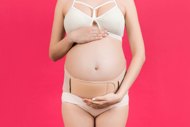 Primer plano de la banda de maternidad elástica vestida en mujer embarazada en ropa interior para reducir el dolor de espalda en el fondo rosa con espacio para copiar. Concepto de cinturón de soporte abdominal ortopédico
. - Foto, imagen