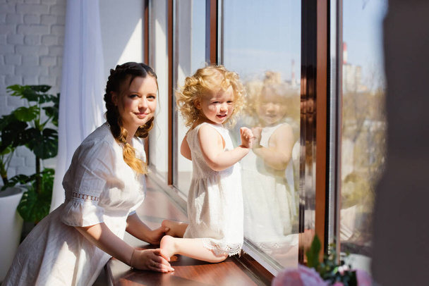 Mother and daughter on the windowsill looking out the window. Happy family at home on the background of a large window.Stay at home. Home Quarantine - Valokuva, kuva