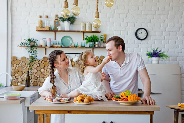 Portrait de famille d'une mère, d'une fille et d'un père heureux posant dans la cuisine pendant le petit déjeuner, mangeant de délicieux macarons, gâteaux, biscuits. De bonnes relations conjugales, l'amour - Photo, image