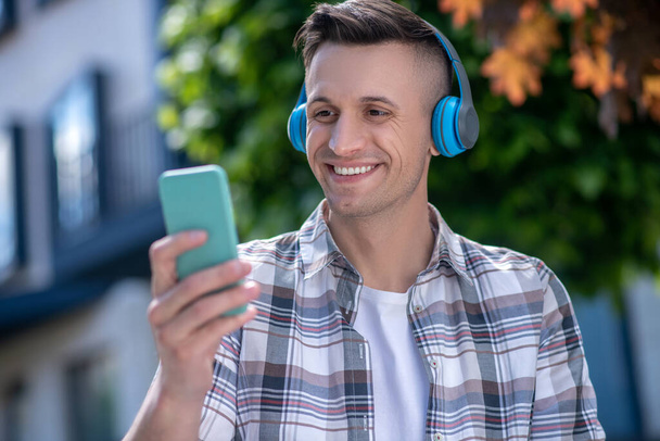 Cheerful dark-haired male wearing headphones, looking at his smartphone - Foto, imagen