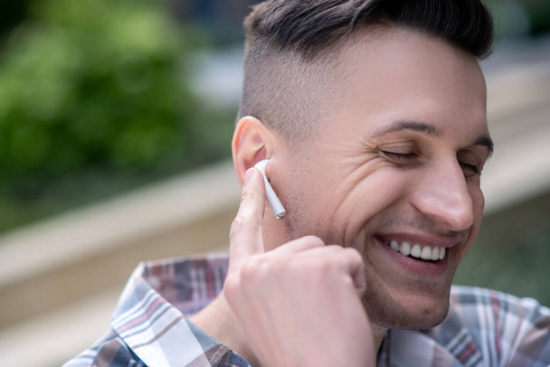 Cheerful dark-haired male wearing wireless earphones, smiling, listening to music - Фото, изображение