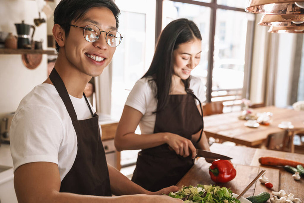 Image of young multicultural couple in aprons laughing and making lunch at cozy kitchen - Foto, Imagem