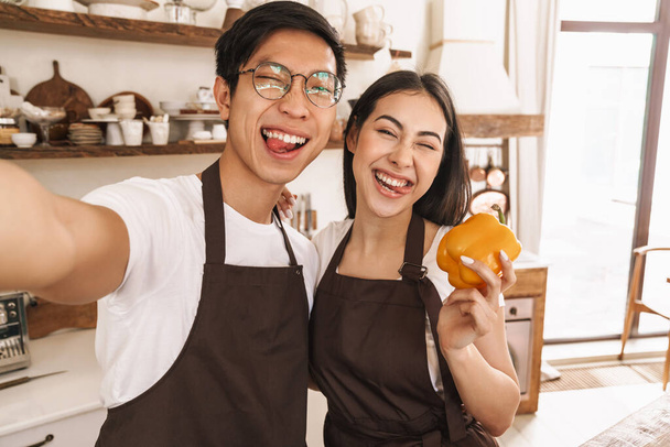 Image of multicultural couple taking selfie on cellphone with sticking out their tongues while holding bell pepper - Foto, Bild