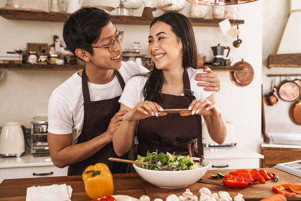 Image of multicultural couple smiling and taking photo on mobile phone while cooking dinner in cozy kitchen - Фото, изображение