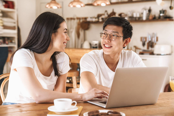 Image of multicultural happy couple smiling and using laptop while sitting at table in cozy kitchen - Φωτογραφία, εικόνα