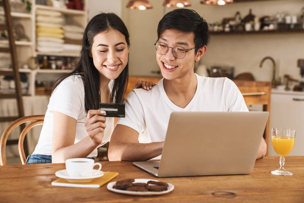 Image of multicultural happy couple holding credit card and using laptop while sitting at table in cozy kitchen - Foto, immagini