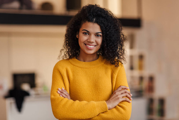 Image of pleased african american woman smiling and looking at camera in living room - Photo, Image
