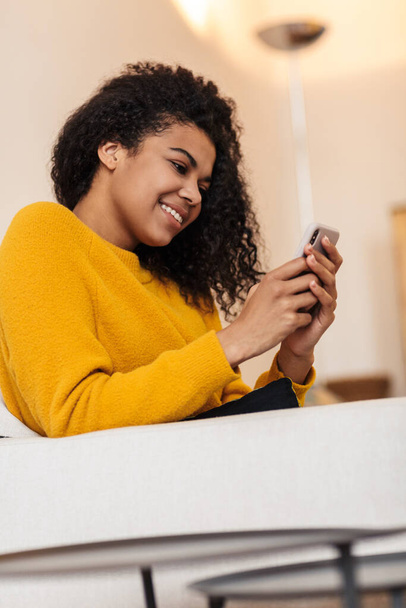 Kuva iloinen afrikkalainen amerikkalainen nainen käyttää matkapuhelinta ja hymyilee istuessaan olohuoneessa
 - Valokuva, kuva