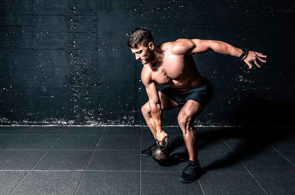 Young strong sweaty focused fit muscular man with big muscles holding heavy kettlebell for cross swing training hard core workout in the gym real people - Foto, Imagem