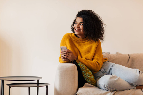 Image of smiling african american woman using cellphone while sitting on couch in living room - Foto, Imagen