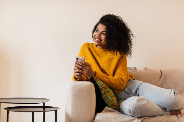 Image of smiling african american woman using cellphone while sitting on couch in living room - Fotoğraf, Görsel