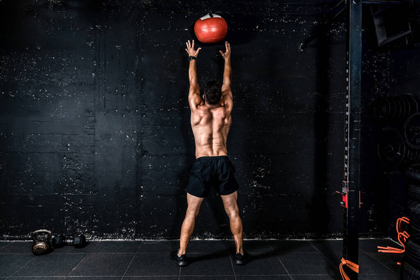 Young strong sweaty focused fit muscular man with big muscles doing throwing medicine ball up on the wall for training hard core workout in the gym real people selective focus - Φωτογραφία, εικόνα