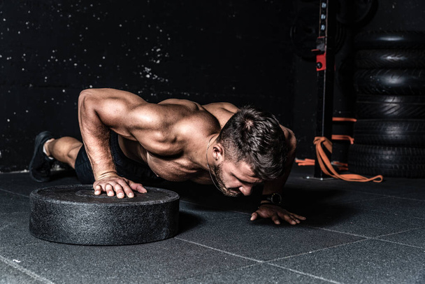Young strong sweaty focused fit muscular man with big muscles performing push ups with one hand on the barbell weight plate for cross training hard core workout in the gym  - Foto, Bild