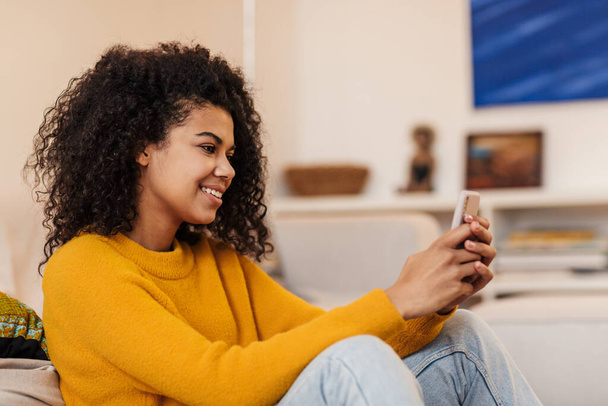 Image of smiling african american woman using cellphone while sitting on floor in living room - Zdjęcie, obraz
