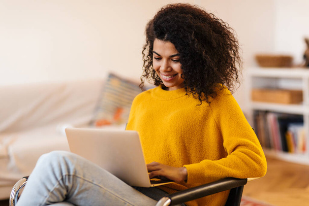 Image of cheerful african american woman using laptop while sitting on chair in living room - Foto, Bild