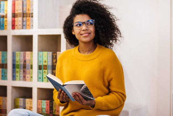 Image of joyful african american woman in eyeglasses reading book while sitting in living room - Photo, image