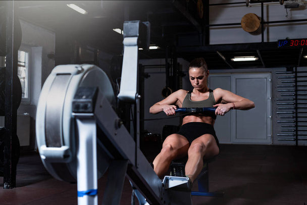 Young strong fit sweaty powerful attractive muscular woman with big muscles doing hard core row heavy cross training workout on indoor rower at the gym real people rowing exercise - Foto, Imagem