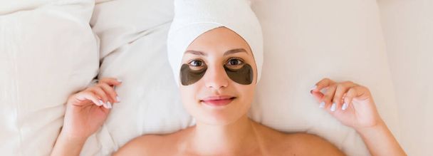 Mujer joven con manchas negras bajo los ojos está acostada y relajada en la cama después de bañarse envuelta en una toalla. Tratamiento de belleza y concepto de cuidado de la piel
. - Foto, imagen