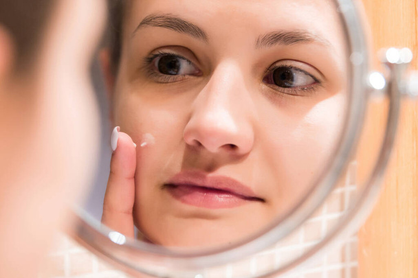 Young girl in front of a bathroom mirror putting cream on a red pimple. Beauty skincare and wellness morning concept. - Photo, Image