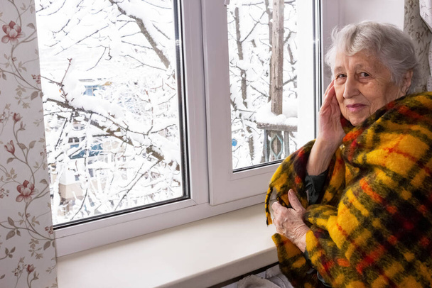 Vieja mujer solitaria sentada cerca de la ventana en su casa y mirando a la cámara y sonriendo en invierno - Foto, Imagen