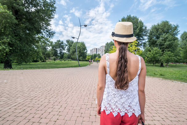 Beautiful young positive and independent French hipster girl walking in the park in the city with hat and sunglasses in retro or vintage fashion clothes view from back on long hair - Fotoğraf, Görsel