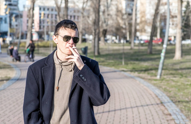 Young casual smoker man with sunglasses in the black coat smoking cigarette outside in the park - Fotografie, Obrázek