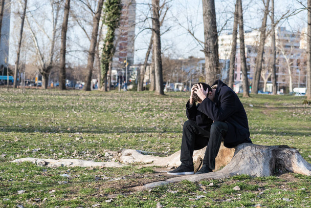 Genç, yalnız, bunalımlı ve endişeli bir adam parkta tek başına oturuyor. Ağlayarak ve düşünerek hayal kırıklığına uğruyor. - Fotoğraf, Görsel