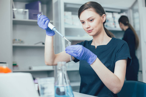 Calm young woman sitting at her workplace in the lab and holding a test tube while measuring the liquid with an automatic pipette dispenser - Zdjęcie, obraz