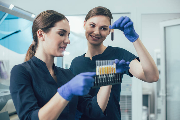 Gladsome scientist holding a test tube rack and her happy colleague taking one blood sample and looking at it with a smile - Photo, image