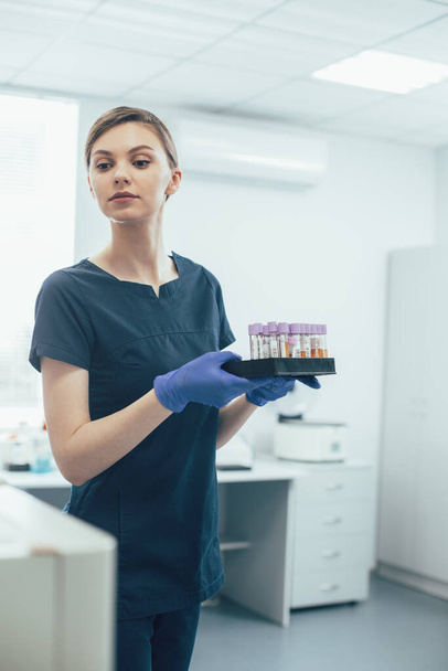 Νεαρή γυναίκα που εργάζεται στο εργαστήριο με δοκιμαστικούς σωλήνες και φοράει λαστιχένια γάντια ενώ κρατά δείγματα - Φωτογραφία, εικόνα