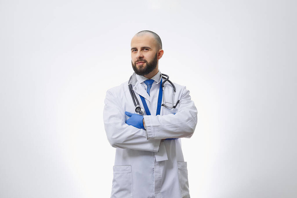 Een arts met armen gekruist op zijn borst in wegwerp medische handschoenen met een stethoscoop. Een kale arts met een baard bereidt zich voor om een patiënt te onderzoeken. - Foto, afbeelding