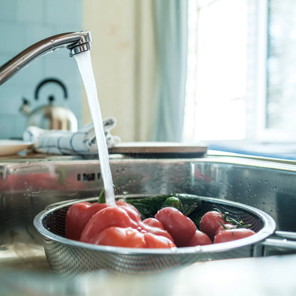Verduras frescas bajo un chorro de agua en un colador en el fregadero, en la cocina, sobre el fondo de una toalla y una tetera
. - Foto, Imagen