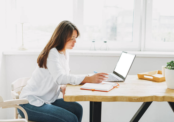 Werken jonge brunette vrouw in wit shirt kleding werken op laptop met wit scherm thuis in helder interieur - Foto, afbeelding