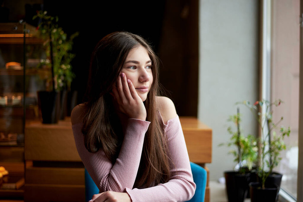 Mladá brunetka, v růžovém svetru, sedí v útulné kavárně se zelenými rostlinami, dívá se na okno v zimě na podzim, přemýšlí. Student si dává pauzu na oběd v kavárně. Nedělní volný čas. - Fotografie, Obrázek