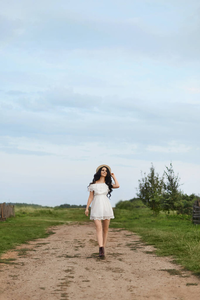 Beautiful girl in white summer dress touching straw hat and smiling while standing on rural road and looking away. Young brunette woman in short white dress walking by a rural road. Summertime - Foto, Imagen