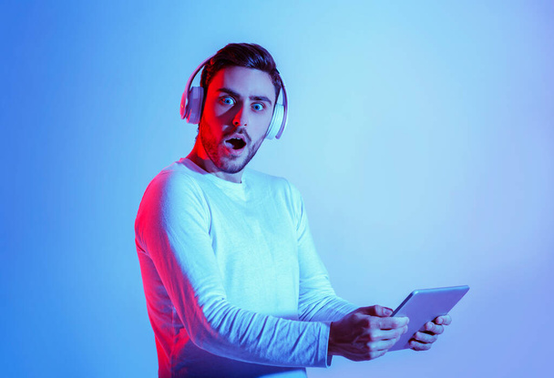 Schokkend concept. Millennial man in hoofdtelefoon met open mond en holding tablet - Foto, afbeelding