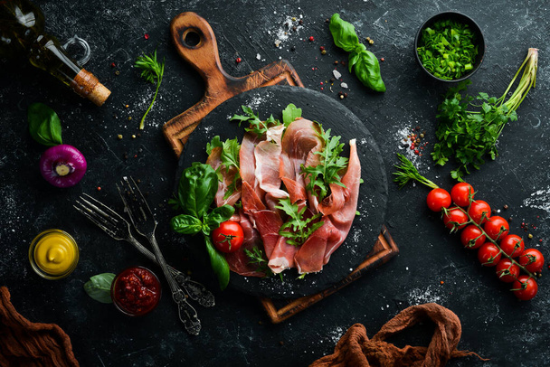 Prosciutto with arugula and basil on a black plate. Spanish cuisine. Top view. Free space for your text. - Φωτογραφία, εικόνα